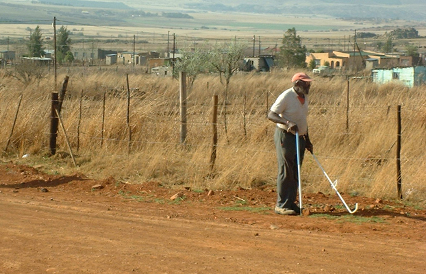Both photos show a man wearing a white T-shirt with a collar, grey slacks, and an orange baseball cap, walking along a dirt road lined with a barbed-wire fence about a high as his shoulder -- on the other side of the fence are fields of yellow grass about as tall as his chest, and beyond that are a half-dozen one-story buildings and green hills beyond them. He uses a white support cane with his right hand, and with his left hand he reaches a long white cane with a bundu basher tip toward the fence.